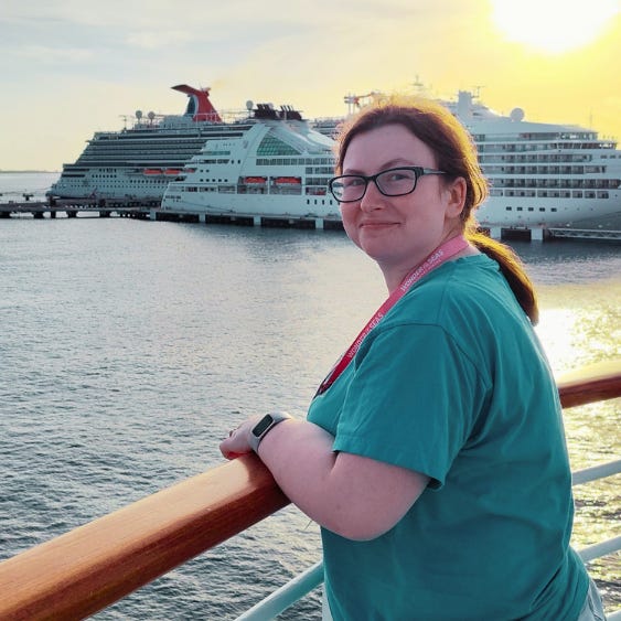 Me, a woman with brown hair in a ponytail looking at the camera while body is facing the ocean (specifically port Cozumel) on the decks of Radiance of the Seas. Behind her is a beautiful sunset and two ships, one carnival and the other unknown. 