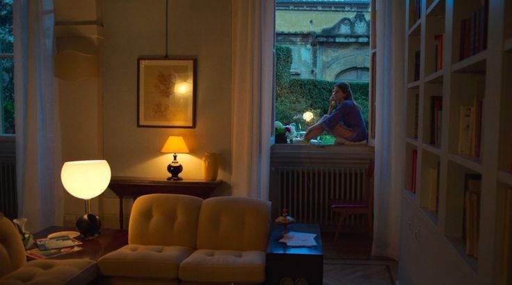 This may contain: a living room filled with furniture next to a window covered in bookshelves and a lamp