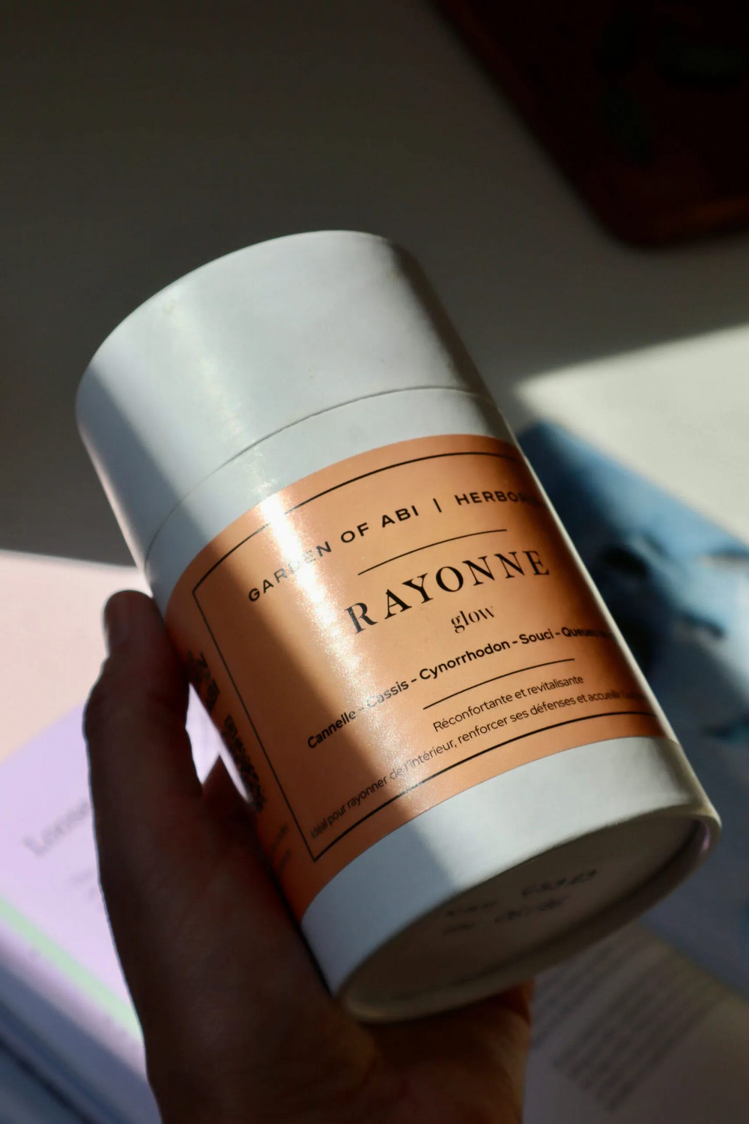 A hand holding a white cylindrical tin of Rayonne tea in soft sunlight. The peach label reads “Garden of Abi”, “Rayonne” and “glow”, with French ingredient and description text. A hand holding a white cylindrical tin of Rayonne tea in soft sunlight. The peach label reads “Garden of Abi”, “Rayonne” and “glow”, with French ingredient and description text.