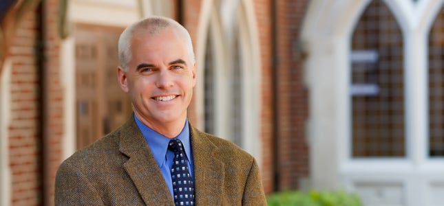 Photo of a smiling middle-aged white man in a suit and tie in front of a brick building.
