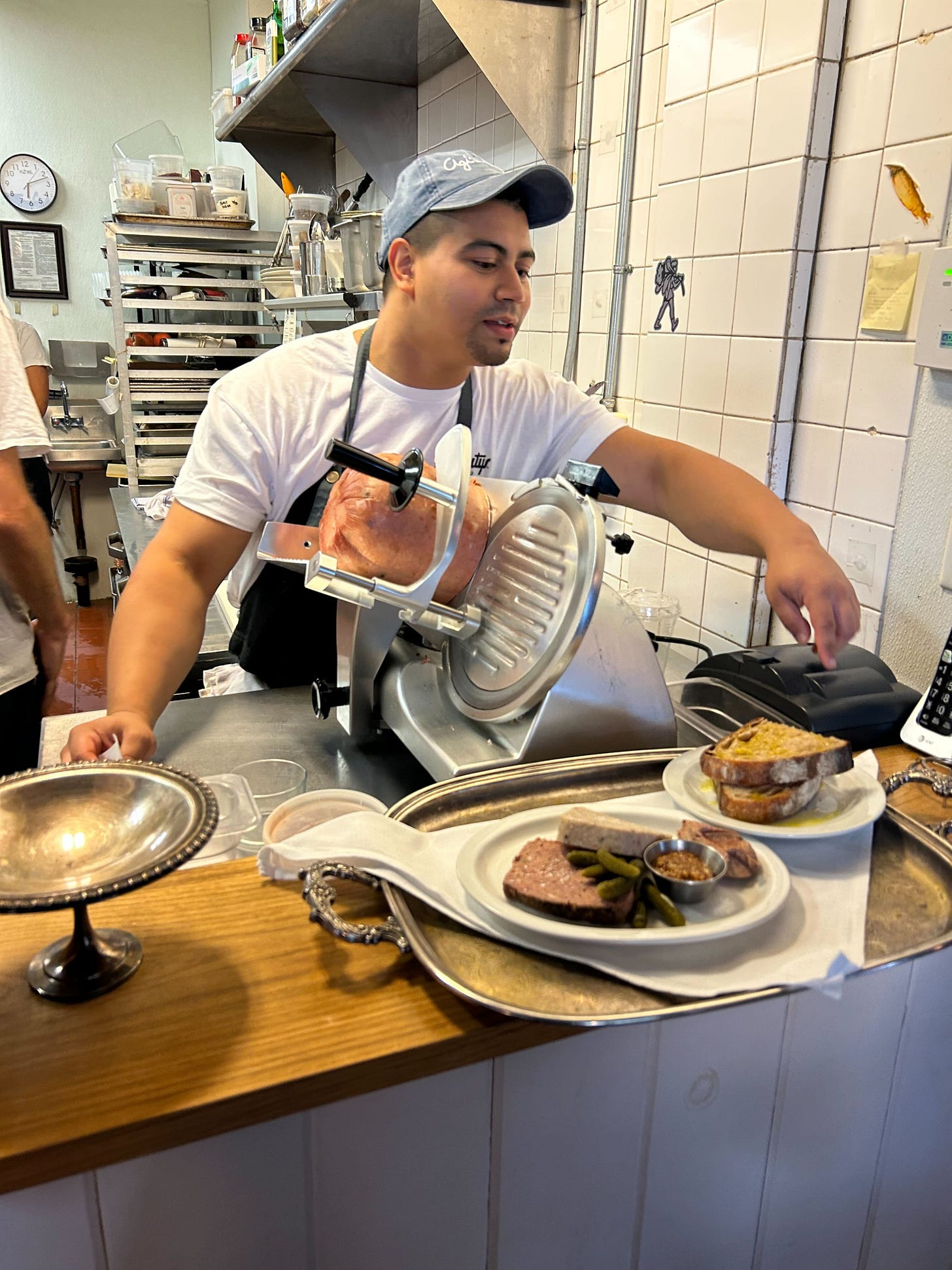 A man in an apron standing in a kitchen in front of a deli-style meat slicer A man in an apron standing in a kitchen in front of a deli-style meat slicer