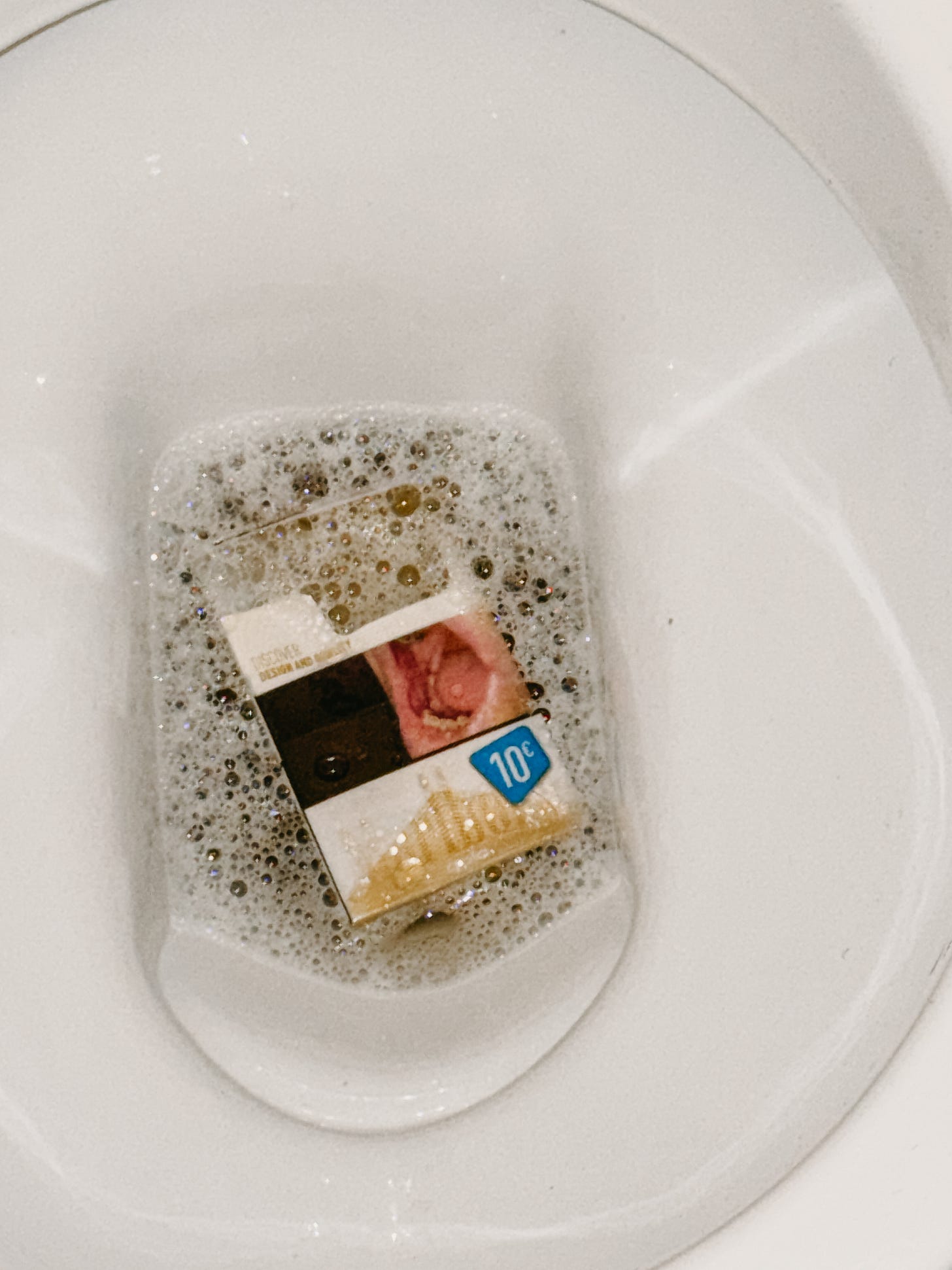 A cigarette carton floating in a toilet bowl filled with soapy bubbles. The carton has a graphic warning image of a diseased mouth and a blue “10¢” price tag. 