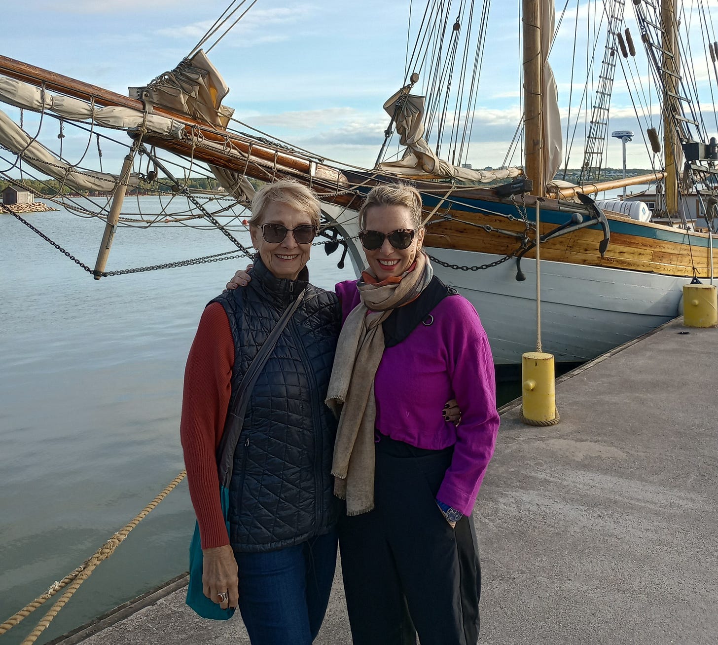 Two blond women dressed for a chilly afternoon, hug one another in front of an old sailing ship. One woman with short hair is wearing a black vest. The other is wearing a pink sweater.