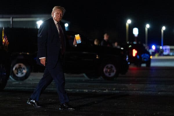 While walking on tarmac at night, President Trump lifts a phone in his left hand so it faces the camera. 