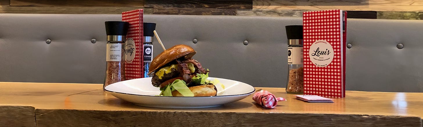 Ein Burger mit Rindfleisch, Käse, Speck und Salat steht auf einem weissen Teller auf einem Holztisch in einem Restaurant. Im Hintergrund sind rote karierte Speisekarten, Gewürzmühlen und eine gepolsterte Sitzbank zu sehen.