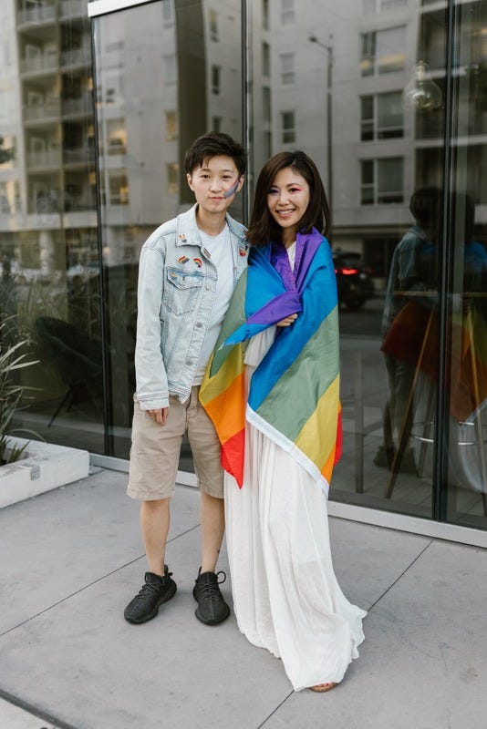 Photo of couple standing together. One person is wearing a rainbow flag around their shoulders. Photo of couple standing together. One person is wearing a rainbow flag around their shoulders.