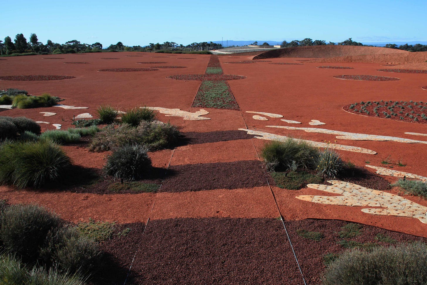 A vast garden mostly with fine red gravel and no plants, with small darker circles and a few plants in the foreground