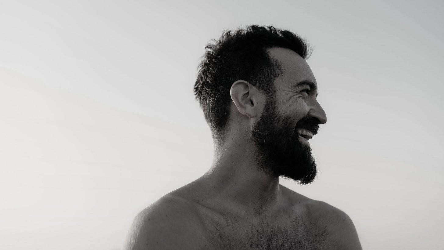 A shirtless and smiling man with a receding hair line looks to the right.