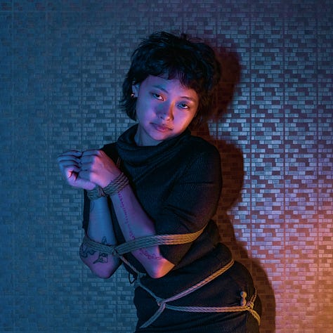 A girl dressed in black is posing with confidence, tied in ropes, over a blue and orange tiled background.