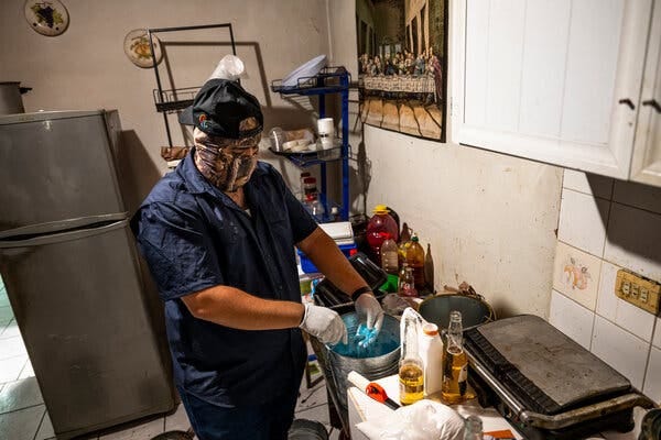 A masked person in a blue jumpsuit mixing drugs in a silver pot in a kitchen. A fridge is in the rear and an image of “The Last Supper” is on a wall.