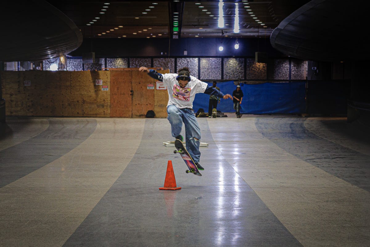 Skateboarder mid-jump over an orange cone in an open indoor space at South Station, surrounded by plywood construction walls. Skateboarder mid-jump over an orange cone in an open indoor space at South Station, surrounded by plywood construction walls.
