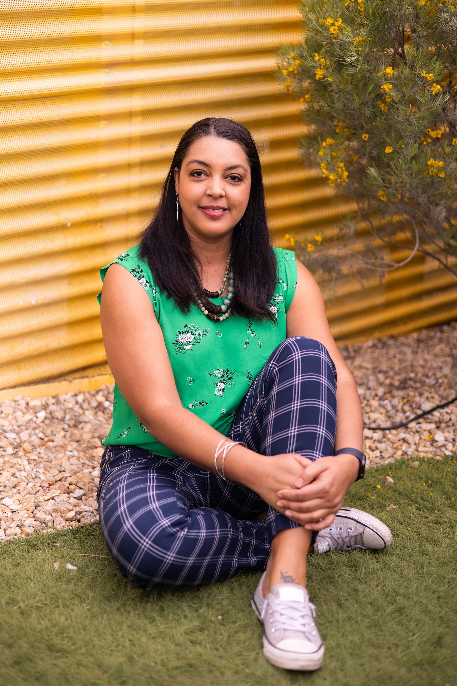 A mixed race woman sits crosslegged on the grass in front of a wavy yellow wall. She is wearing a green top, blue pants and gray Chucks. 