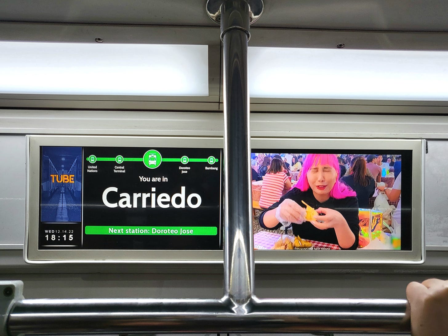 A passenger information screen mounted to the side wall of an LRTA Line 1 train. The screen contains: (Left) the TUBE logo along with the date and time; (Center) a five-station strip map, the current station as "Carriedo", and the next station as "Doroteo Jose"; (Right) a widescreen-format video ad