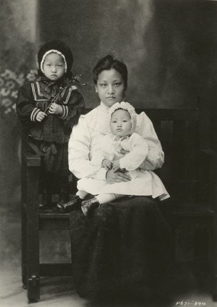 Little Anna dressed in a white dress and bonnet sits in her mother’s lap, while her older sister Lulu stands next to them on the bench, circa 1905 Little Anna dressed in a white dress and bonnet sits in her mother’s lap, while her older sister Lulu stands next to them on the bench, circa 1905