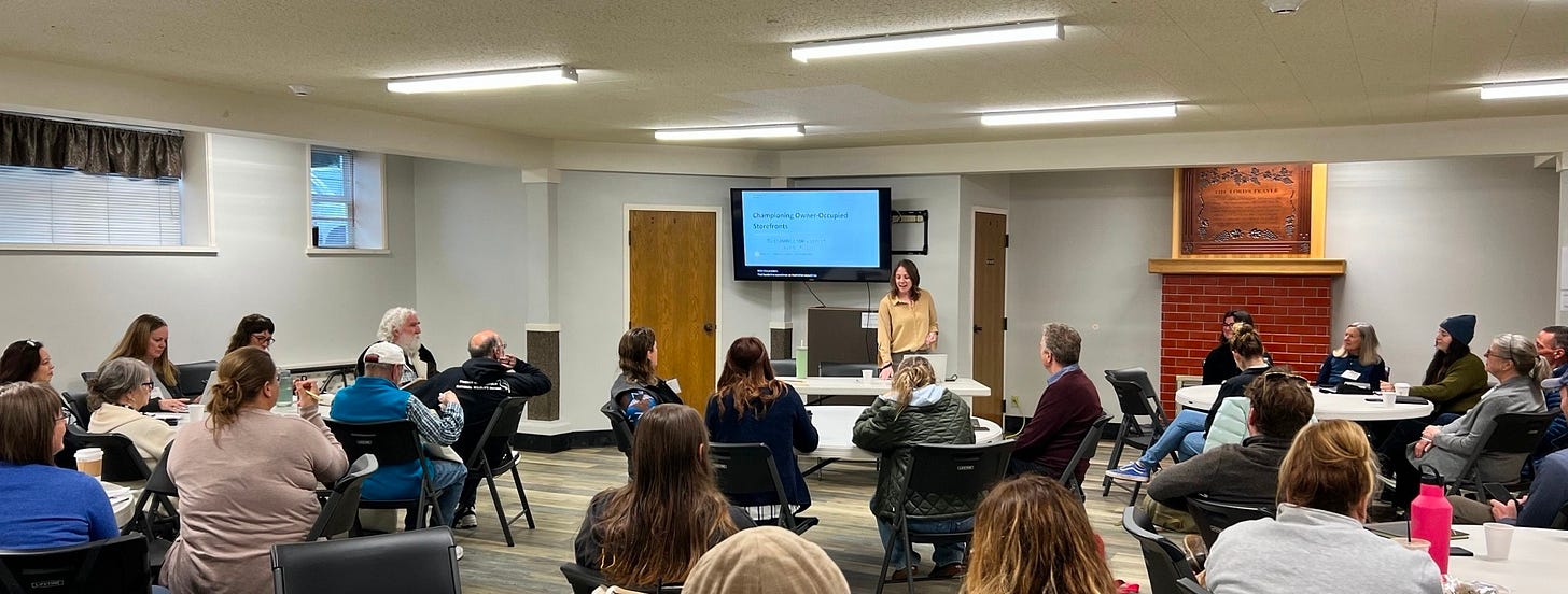 person presenting at the front of a room full of people with a television behind them