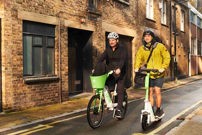 Two men are travelling along street - one is on a white and green branded Lime bicycle and the other on a Lime e-scooter Two men are travelling along street - one is on a white and green branded Lime bicycle and the other on a Lime e-scooter