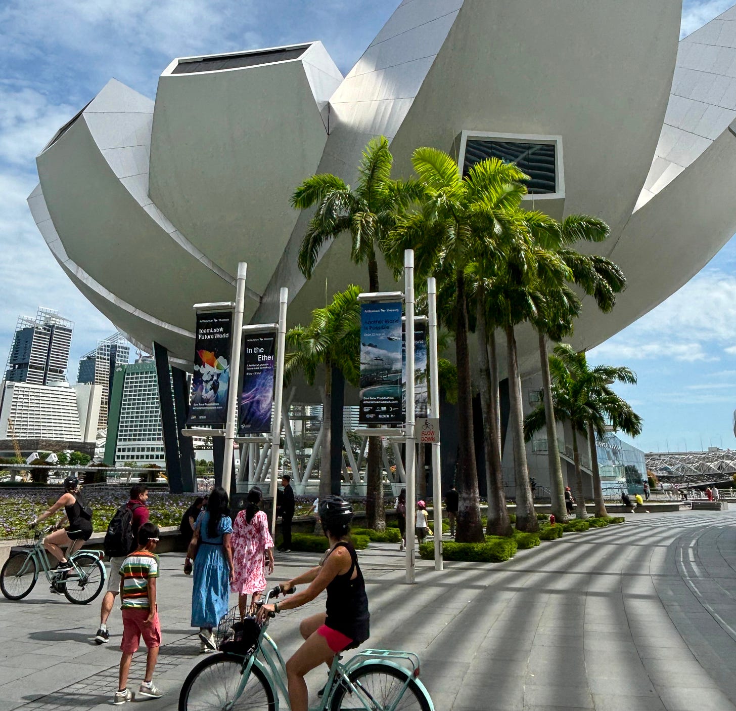 A futuristic looking building surrounded by palm trees. In the foreground is a cyclist and some pedestrians