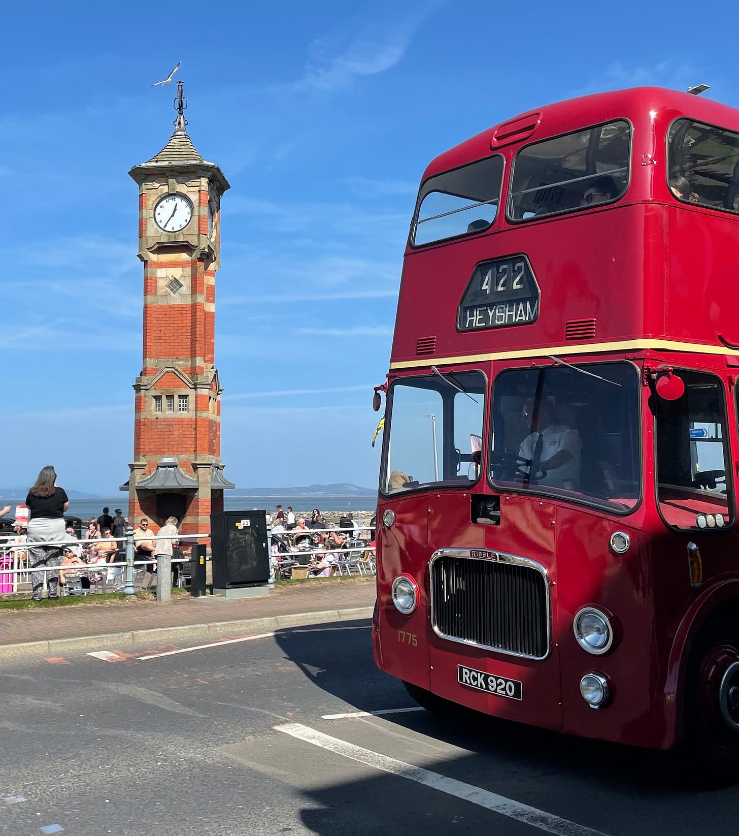 A bright red double-decker vintage bus in front of a clock tower, in the sunshine
