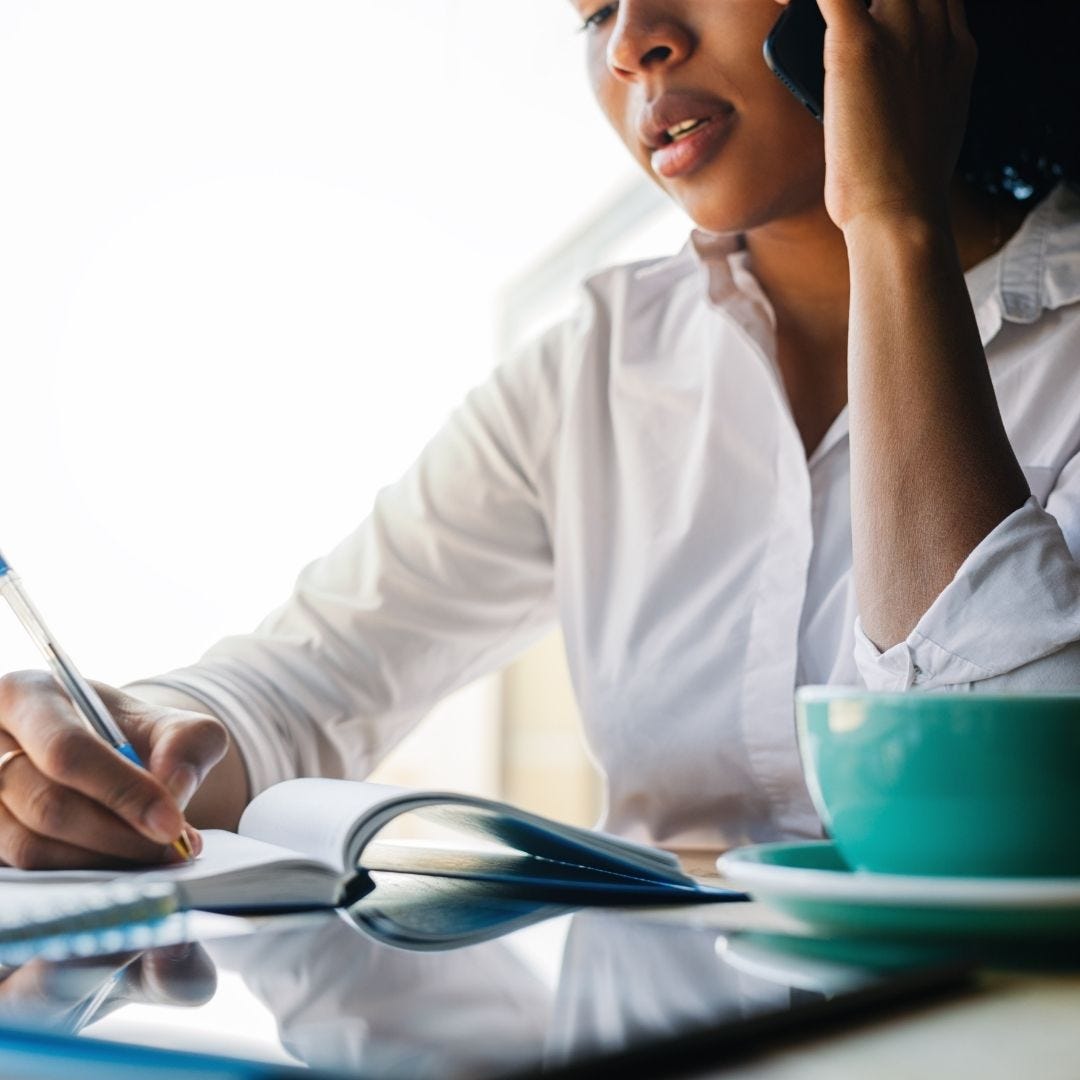 Woman taking handwritten notes on a notepad while holding a cell phone to her ear. A tablet and coffee mug rest on the table.