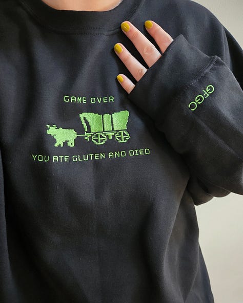 Modeling merch: A hot pink sweatshirt with white embroidered text on the front chest that says "gluten gives me the ick" and "GFGC" on the left sleeve near the wrist. A vintage wash denim baseball cap with an embroidered white pixelated chat bubble and black "GFGC" inside. A black sweatshirt with a green embroidered design on the front chest that says "GAME OVER, YOU ATE GLUTEN AND DIED" with a pixelated ox and wagon and "GFGC" on the left sleeve near the wrist.