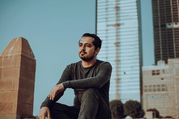 A man in black clothes staring off into the distance with corporate buildings in the background
