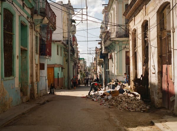People in Havana walking on a street with brightly painted buildings and piles of debris and garbage on the sidewalk.