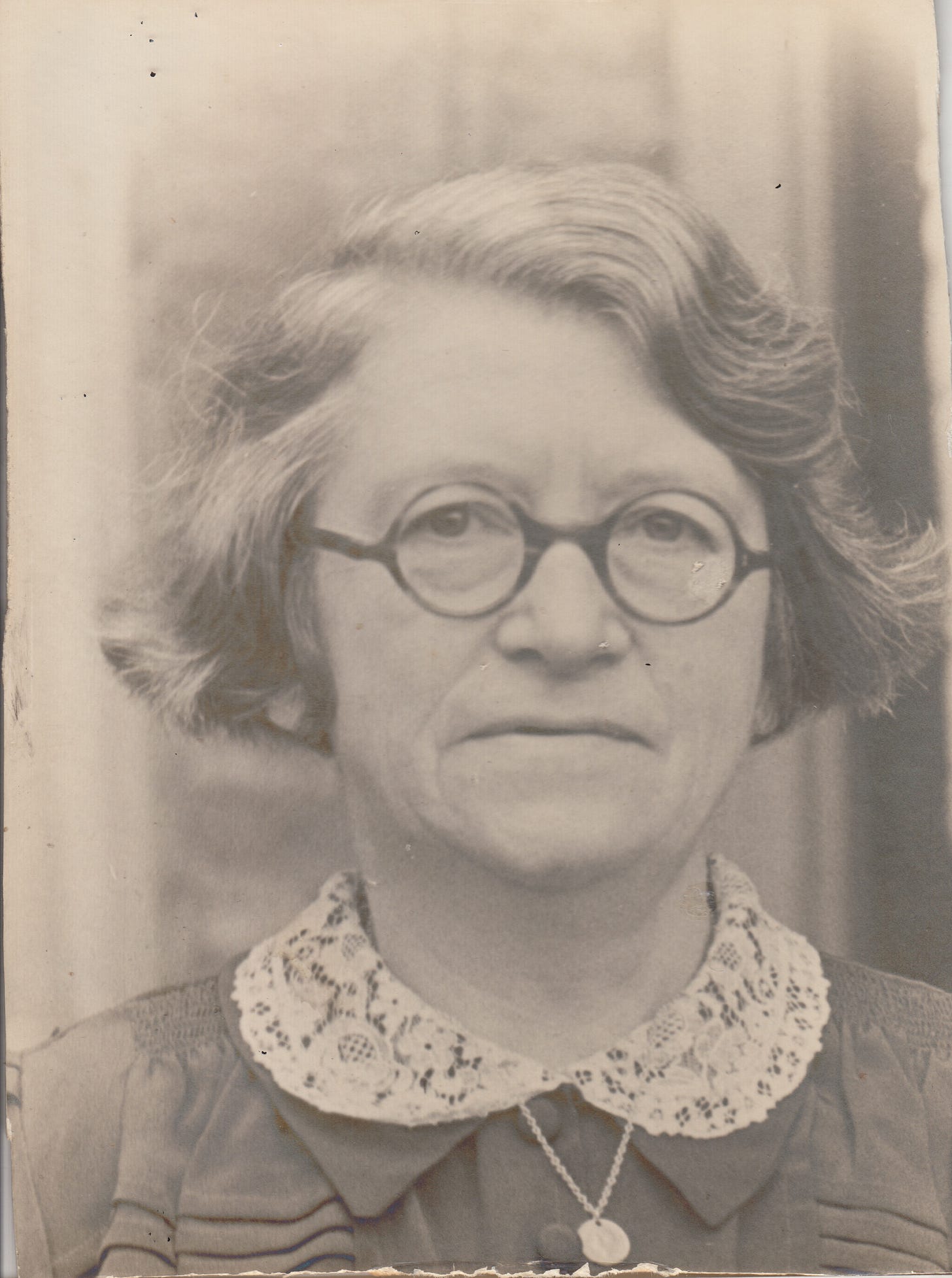 A sepia/black and white photographic portrait of a late middle-aged woman with thick hair, round glasses, and a stern expression on her face. She has a lace collar on the neck of her dress.