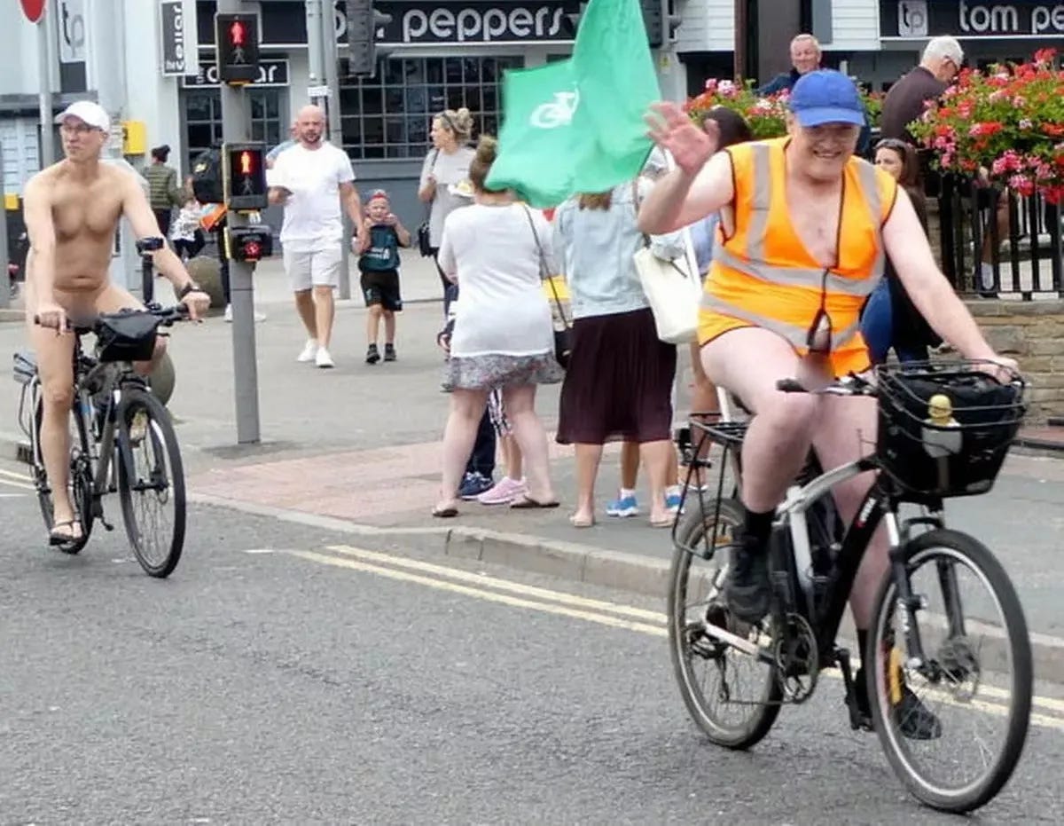 Two cyclists ride through a busy intersection during a World Naked Bike Ride. One wears only a cap and sandals, while the other sports a hi-vis vest and a smile, waving to onlookers. Pedestrians and families watch from the sidewalk.