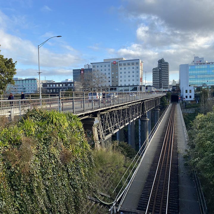 Left: A photograph from the footpath, looking west, showing footpath on both sides, with two lanes of traffic which doubles as sharrows. Right: The view from the Sonny Carpark footbridge, looking down onto the rail bridge, and then across and over onto Claudelands Bridge.
