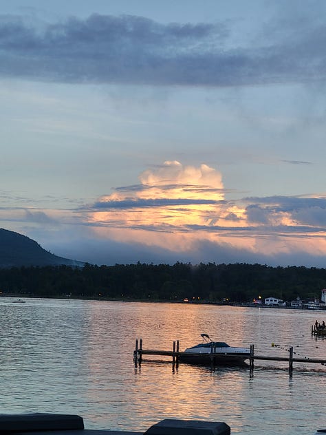 A collection of 9 images depicting: the shoreline of Lake George including a lighthouse and steamboat; a beautiful sunset with glowing pink clouds over the lake, being photographed from a deck behind a woman with long, wet hair who is photographing the same clouds; the lake on a sunny day with water sparkling in front of low mountains; a duck looking at the camera from a sandy beach; a young teenage boy sitting at a table with a small town street in the background; the same teenage boy wearing a wet swimsuit, sitting astride a bear statue at an indoor water park; the same teenage boy sitting with his feet up in his chair on the deck by the lake, wearing a coat and headphones as he stairs at his phone; a street view of the small downtown area with mountains rising in the background; pink, fluffy, glowing clouds over the lake at sunset.