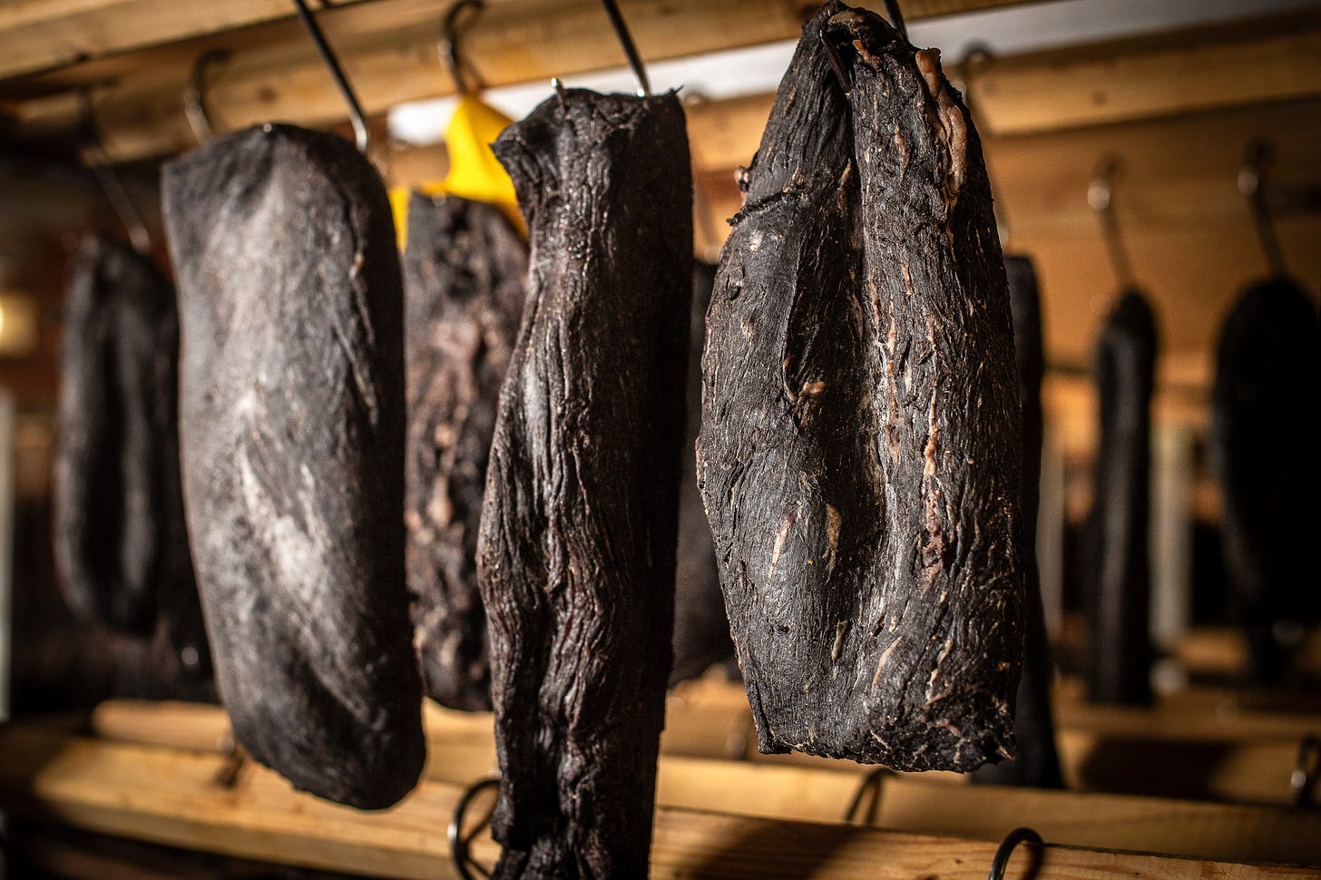 In der Fleischtrocknerei Sialm hängen mehrere dunkle, längliche Stücke Trockenfleisch an Haken von einer Holzstange. Die Oberfläche ist runzlig und trocken, das Licht betont die Struktur des gereiften Fleisches. Im Hintergrund sind weitere Fleischstücke unscharf erkennbar.