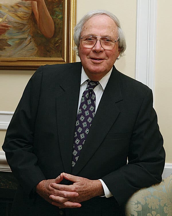 A photo of a round-faced man with somewhat long but thinning gray hair that covers his ears. He wears metal-framed eyeglasses and a dark suit and dark patterned necktie over a white shirt. His hands are folded in front of him as he leans against a high-backed upholstered chair, one elbow resting on it. 