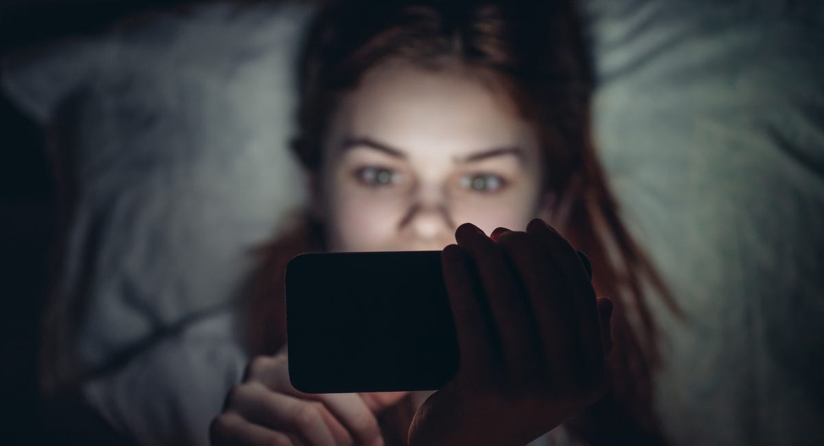 Vrouw ligt in bed in het donker en kijkt naar een smartphone die haar gezicht verlicht.