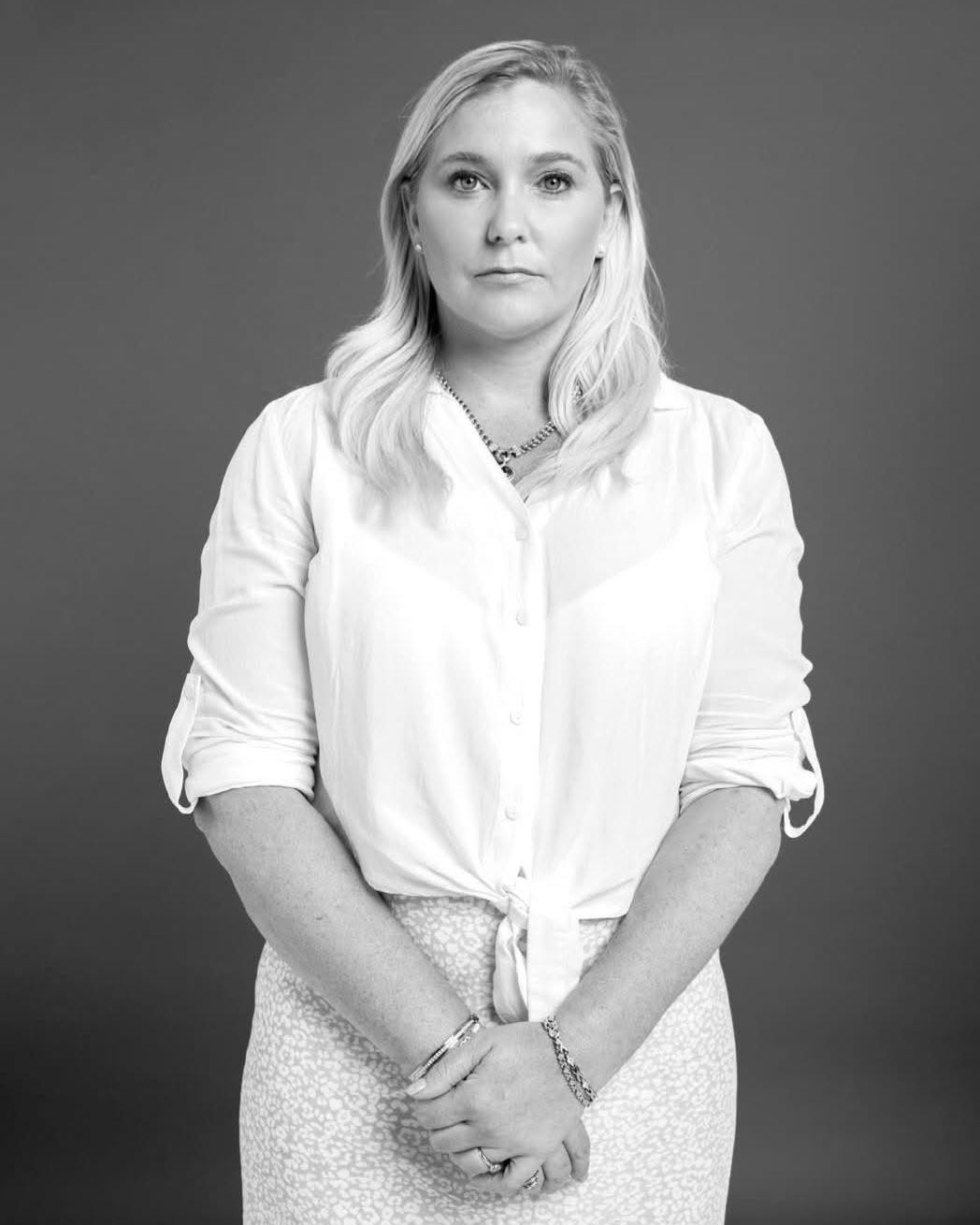 A black-and-white photo of Virginia Giuffre, standing calmly in a white blouse and looking straight at the camera with quiet strength.
