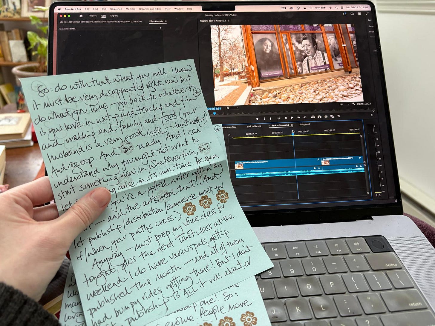 We see a hand holding a letter written on two blue pieces of paper in beautiful, loopy handwriting. Behind them is a computer. On the screen is an Adobe Premiere Pro timeline with an image of a wall with two portraits on them, one of Bobbie Louise Hawkins and one of Chögyam Trungpa Rinpoche. It is daytime. We see a hand holding a letter written on two blue pieces of paper in beautiful, loopy handwriting. Behind them is a computer. On the screen is an Adobe Premiere Pro timeline with an image of a wall with two portraits on them, one of Bobbie Louise Hawkins and one of Chögyam Trungpa Rinpoche. It is daytime.