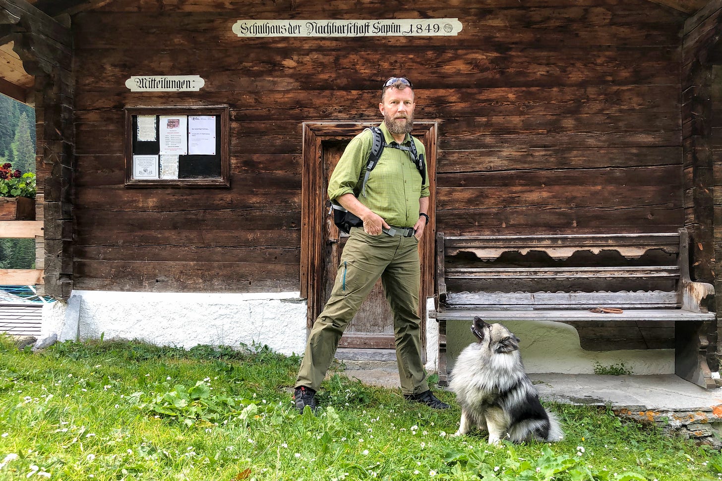 Der Agrarjournalist Jürg Vollmer mit Rucksack steht vor dem historischen Schulhaus von 1849 in der Walsersiedlung Sapün. Neben ihm sitzt sein silbergrauer Wolfsspitz im Gras und blickt aufmerksam zu seinem Besitzer auf. Das aus Holz gebaute Schulhaus diente ab 1873 als Sennerei und ab 1963 als Dorfwäscherei. Heute ist es Teil der malerischen, unter Denkmalschutz stehenden Walsersiedlung Sapün.