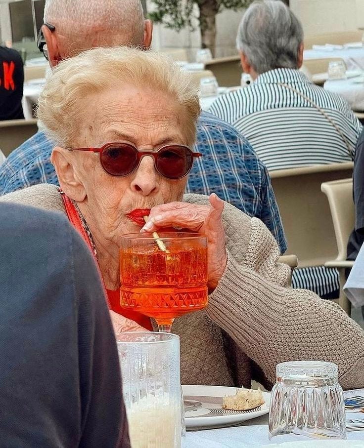 This may contain: an older woman sitting at a table with a drink in her hand