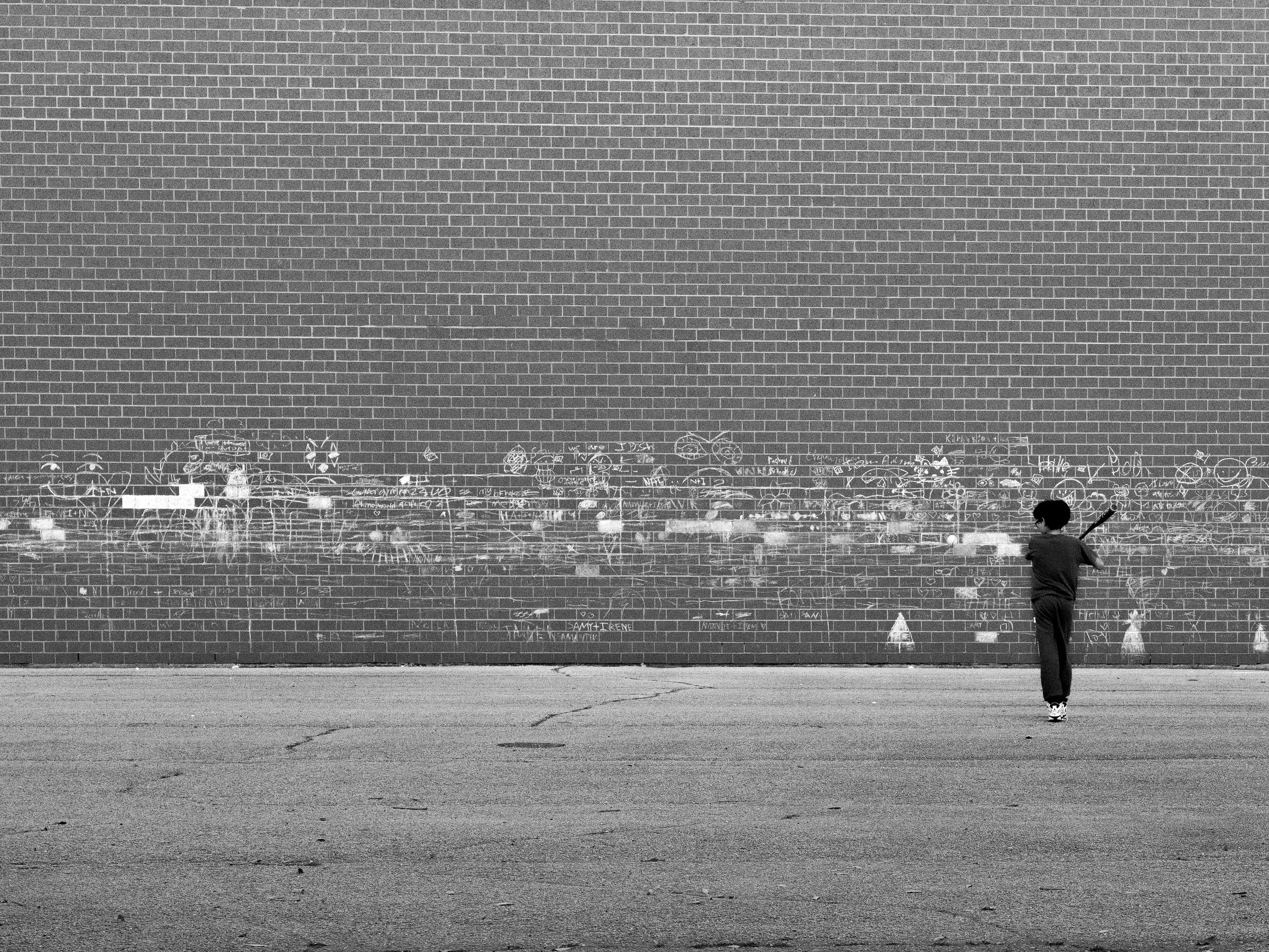Black-and-white photo of a child playing baseball in a concrete school lot. Chalk drawings are in the background on brick wall.