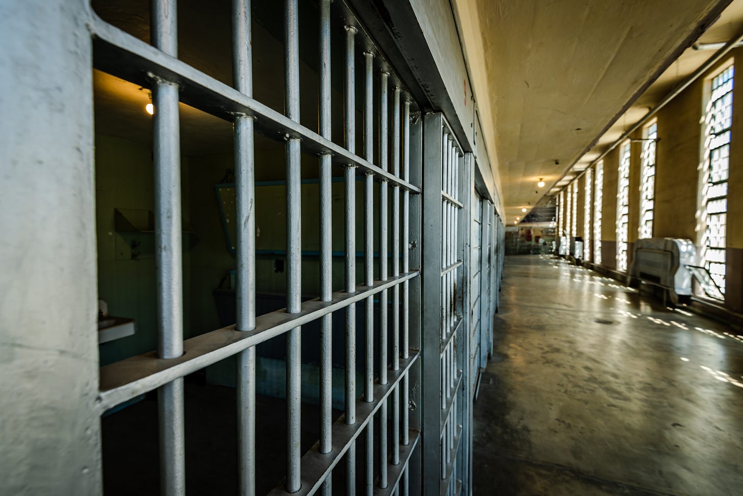 A row of grey prison cell bars looking out on a hallway of windows