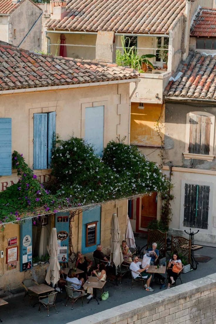 This may contain: several people sitting at outdoor tables in front of an old building with blue shutters