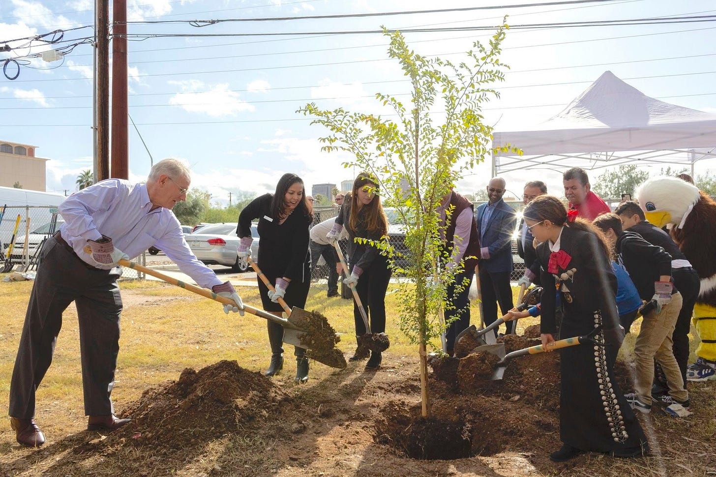 Color photo of a group of adults and middle-school students shoveling soil onto a freshly-placed tree in a hole outside a middle school. It's one of those semi-staged photos where each shovel has only a bit of soil so all four kids, seven adults, and the school mascot, in a eagle costume, can all contribute to planting the tree. The girl in the front of the group of kids is wearing a black charro dress with red bow and silver accents. The school has an acclaimed mariachi music program. 