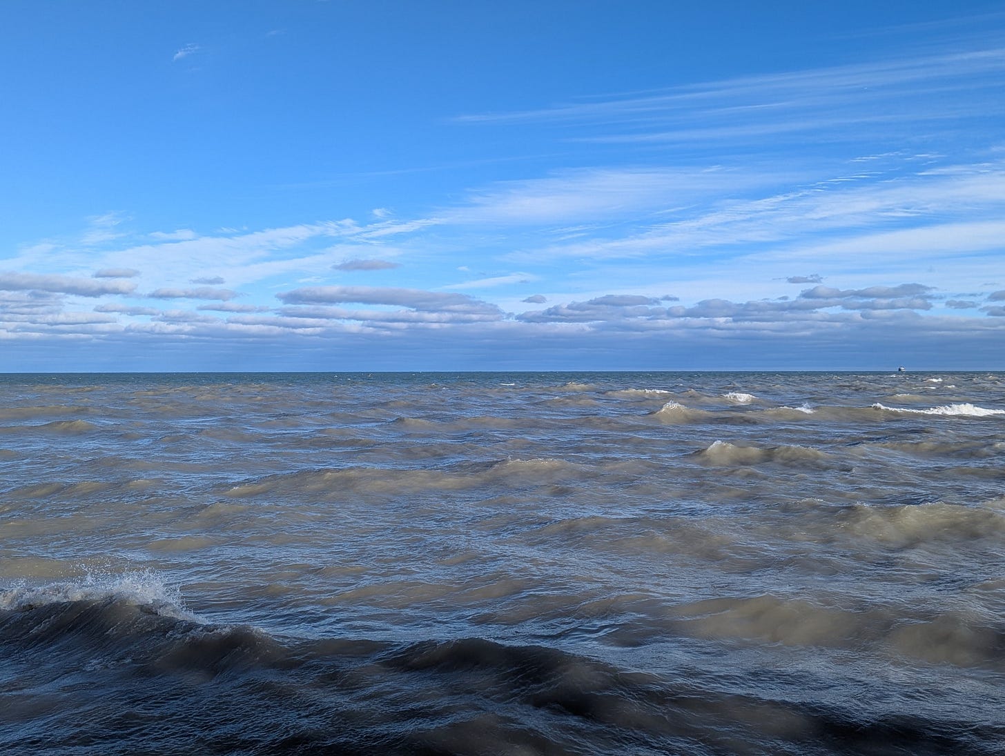A photo of Lake Michigan. Half the image is taken up by a bright blue sky splattered with clouds. Underneath, a grey mass of water churns with uneven waves. There's a shadow along the foreground of the lake, and a strip of greenish blue along the horizon. A photo of Lake Michigan. Half the image is taken up by a bright blue sky splattered with clouds. Underneath, a grey mass of water churns with uneven waves. There's a shadow along the foreground of the lake, and a strip of greenish blue along the horizon.