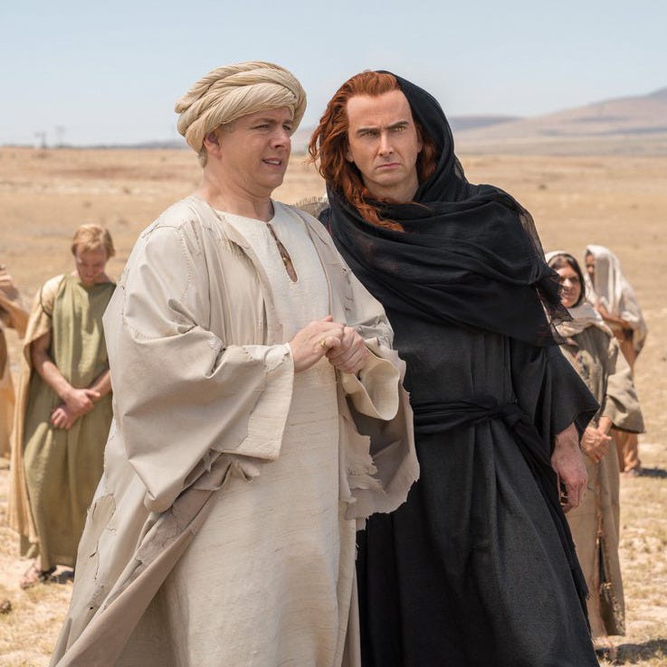 A photo of Aziraphale and Crowley standing in side-by-side in the desert. Aziraphale is dressed in all white with a turban wrapped around his head. Crowley is dressed in all black, her red hair is long, and she has a scarf wrapped around her shoulders and worn over her hair.