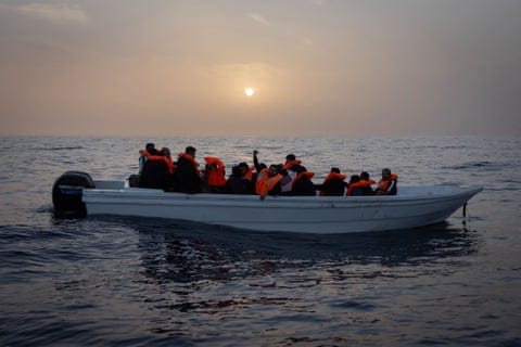 Migrants from Syria and Bangladesh wearing orange life vests in a small boat in the Mediterranean.