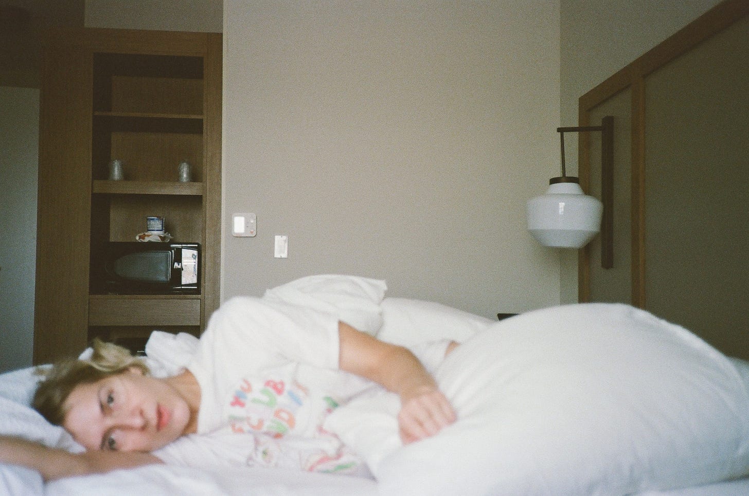 Rebekah lies on her side in a hotel bed, looking toward the camera. She’s surrounded by white pillows and sheets. She is still, nearly expressionless. Behind her, a sterile, mostly empty hotel room. 