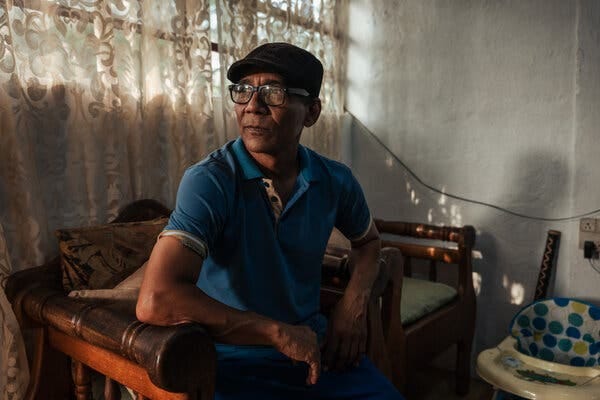 Una persona con un polo azul, gorra oscura y gafas está sentada en un interior. La luz se cuela a través de unas cortinas estampadas detrás de ellos, y se ve un andador de bebé.