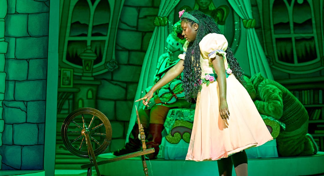 A young woman is standing looking down with her finger outstretched towards a spinning wheel. Her facial expression is blank, as if in a trance. She is wearing bright pink shoes and a light pink dress, with roses around her waistband and in her hair. The backdrop depicts the inside of the castle, with a bed on stage.