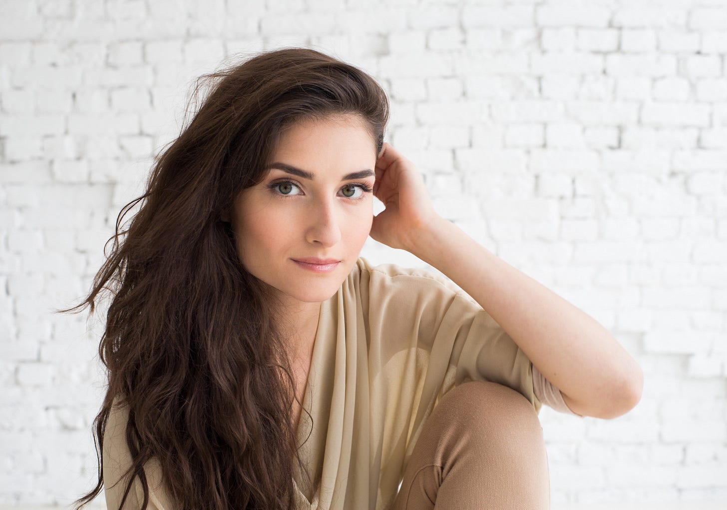 A woman with green eyes and long brown hair tucks her hair behind one ear.