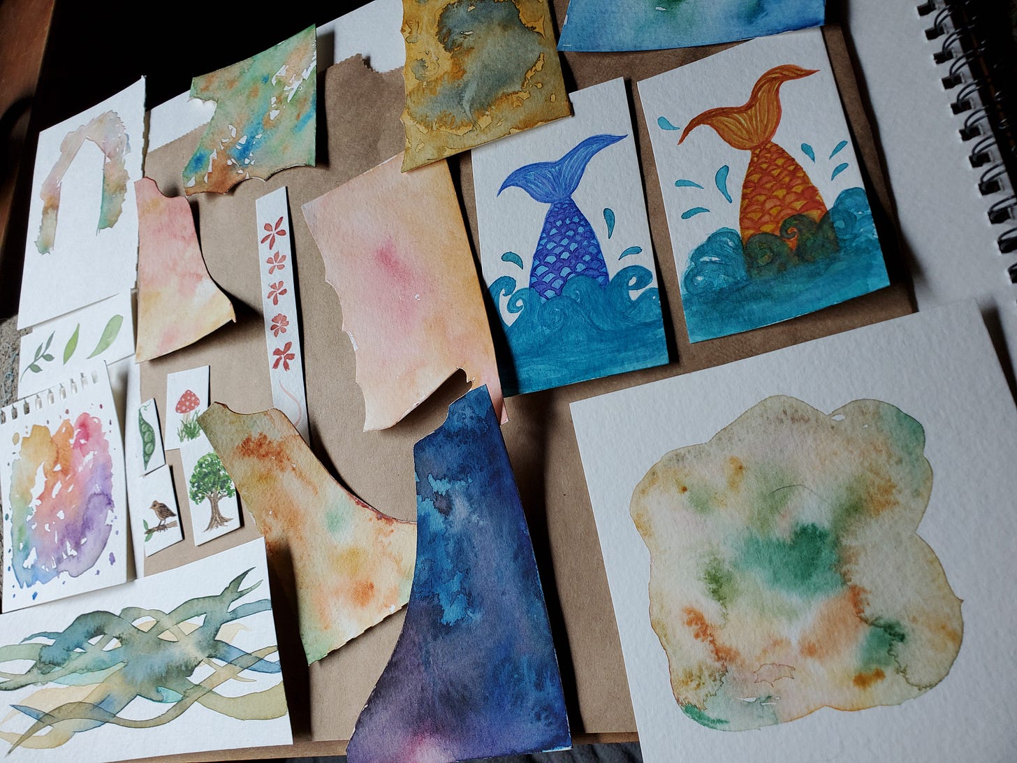 A spread of lots of small pieces of watercolour paper with a variety of different paintings on them. There are a few tiny paintings (a mushroom, a pea pod, a tree, and a bird on a branch), a couple of mermaid tails coming out of the water, and some random patterns and colour mixtures on scraps.