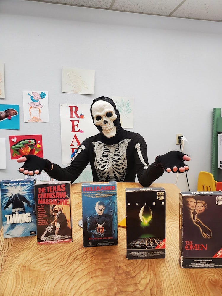 A person in a full-body skeleton suit in mask gestures with open arms at a collection of horror films on VHS tape in front of a wall of children's art and a banner that says "READ".